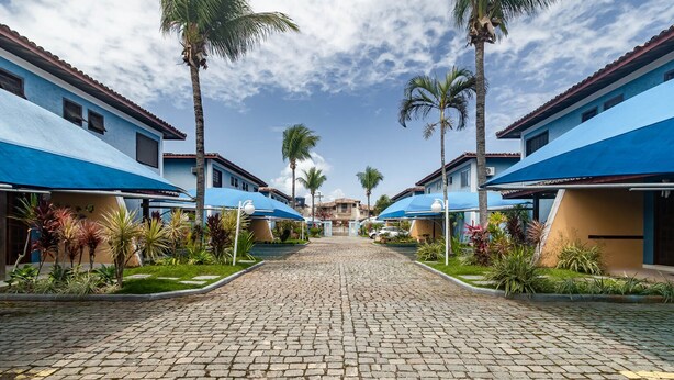 Un hôtel avec un chemin pavé menant à un bâtiment central, bordé de palmiers et flanqué de constructions aux toits bleus.