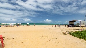 Une plage avec des parasols blancs et des chaises longues, un filet de volley-ball et un petit bâtiment.