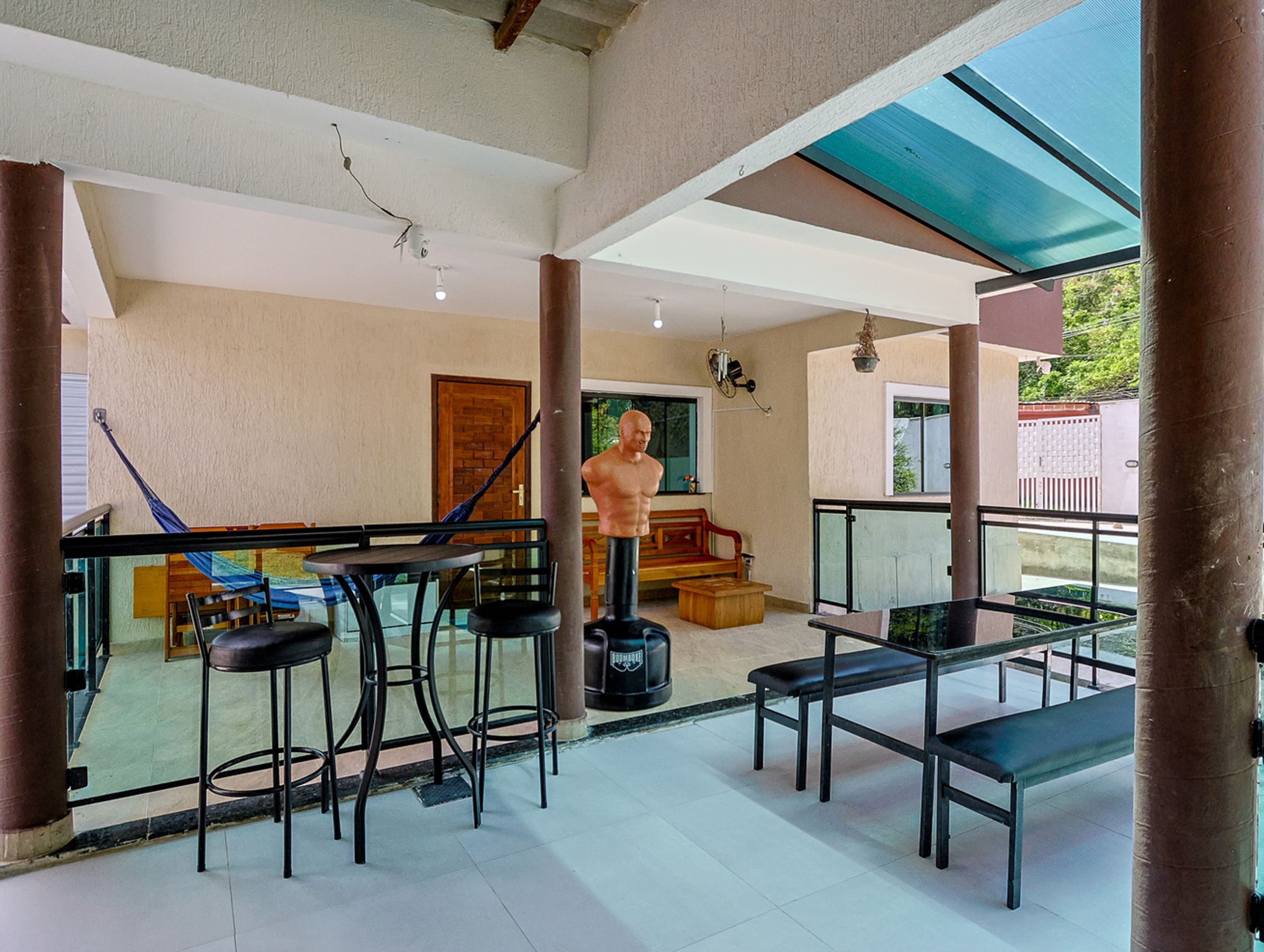 Uma varanda ampla com guarda-corpo de vidro, uma escultura e um espaço de estar com mesa e cadeiras.