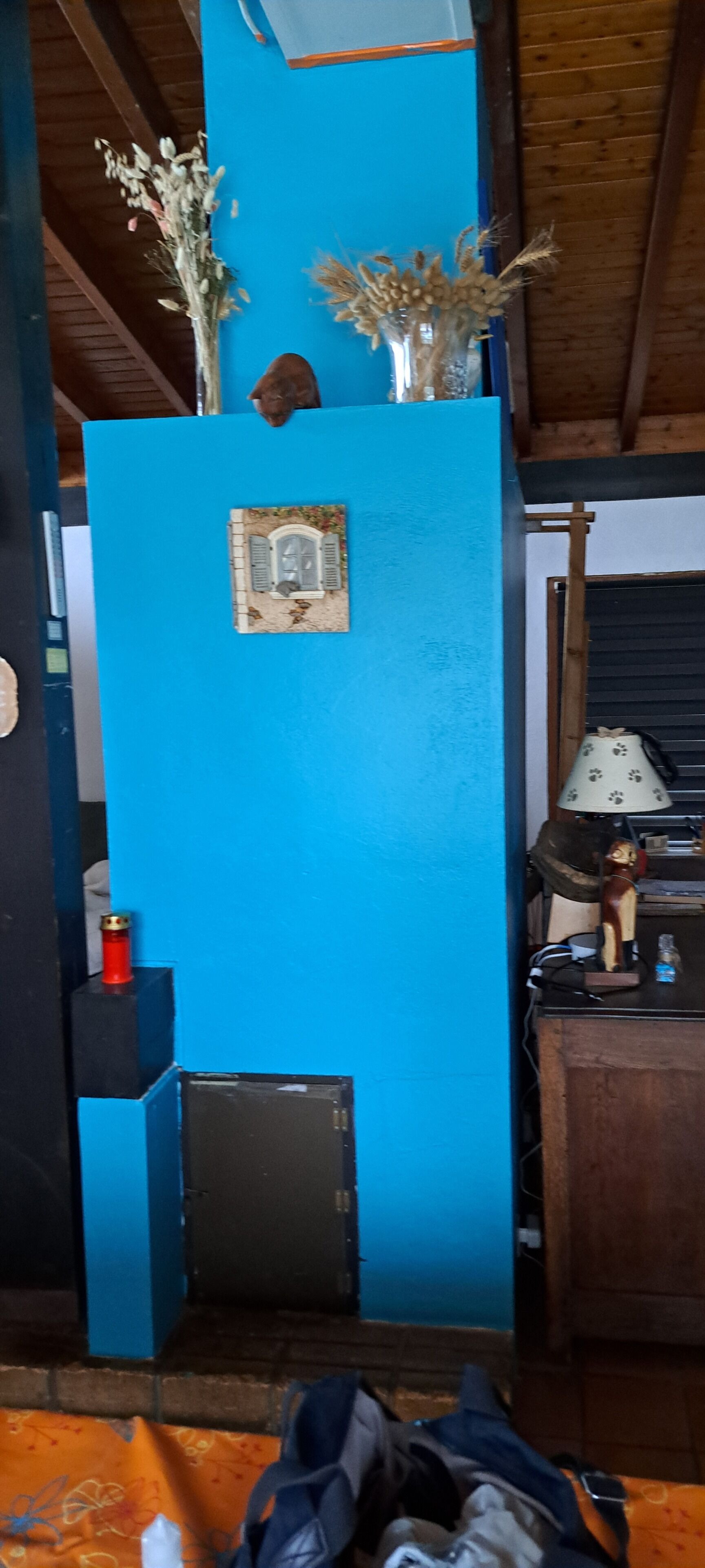 Ein Zimmer mit blauer Wand, einem kleinen Fenster mit Fensterläden, einer Holztür, einer Lampe und einem kleinen Tisch mit einer Flasche.