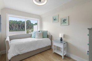 A bedroom with a bed, a nightstand, a window with a view of palm trees, and two framed pictures on the wall. - Villa Bleu 1 (New Smyrna Beach)