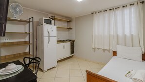 Une chambre d’hôtel compacte comprenant un coin cuisine, un lit avec un oreiller blanc, une table à manger avec des chaises et un ventilateur fixé au mur.