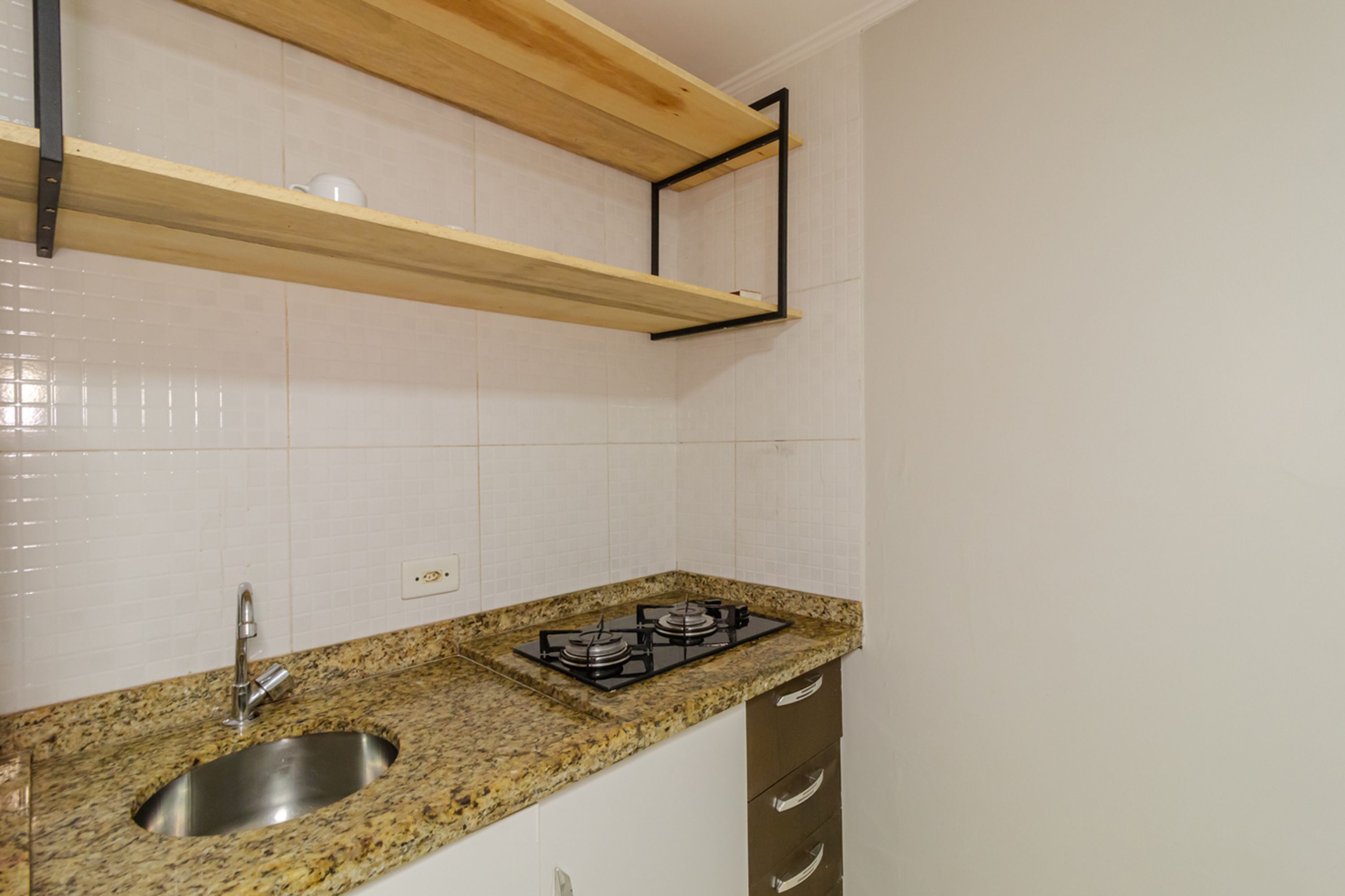 A kitchen with a granite countertop, stainless steel sink, and a gas stove. There are wooden shelves above the countertop and a white tiled backsplash.