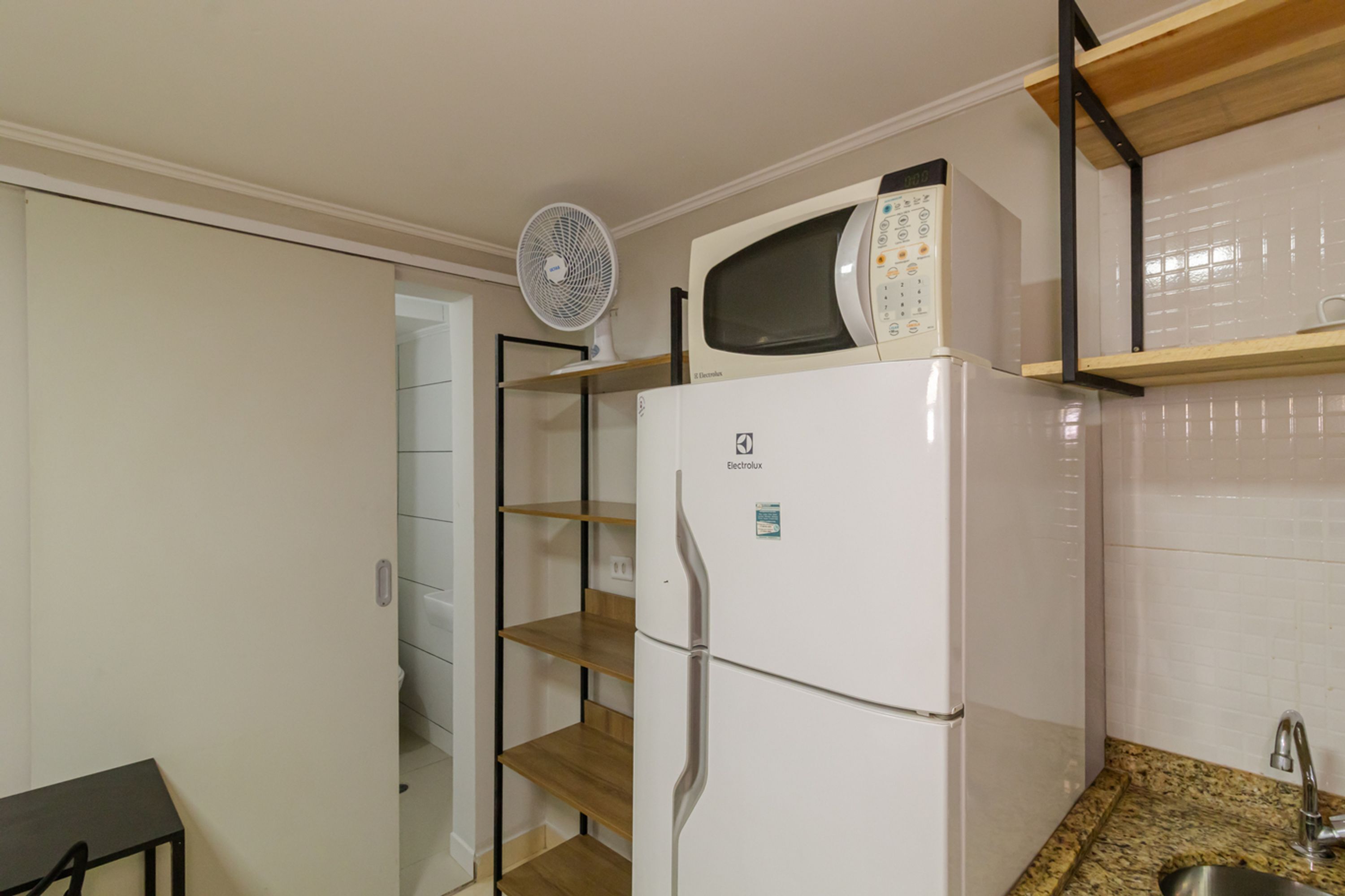 A compact kitchen with a built-in microwave, a refrigerator, and a fan on a shelf.