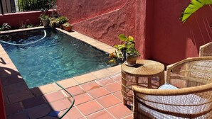 Piscina con parete rossa e recinzione metallica, circondata da piante in vaso e mobili in vimini.