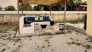 Uma área externa com mobiliário de jardim, incluindo um alpendre coberto, um sofá, duas mesinhas laterais e um aparelho de som.