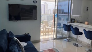 Ein modernes Wohnzimmer mit einem großen Flachbildfernseher, einer Couch und einer Glastür, die auf einen Balkon mit Blick auf die Stadt führt.