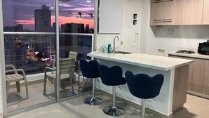 Eine moderne Küche mit einem großen Fenster, einer Bar mit Hockern und Blick auf eine Stadtlandschaft bei Sonnenuntergang.