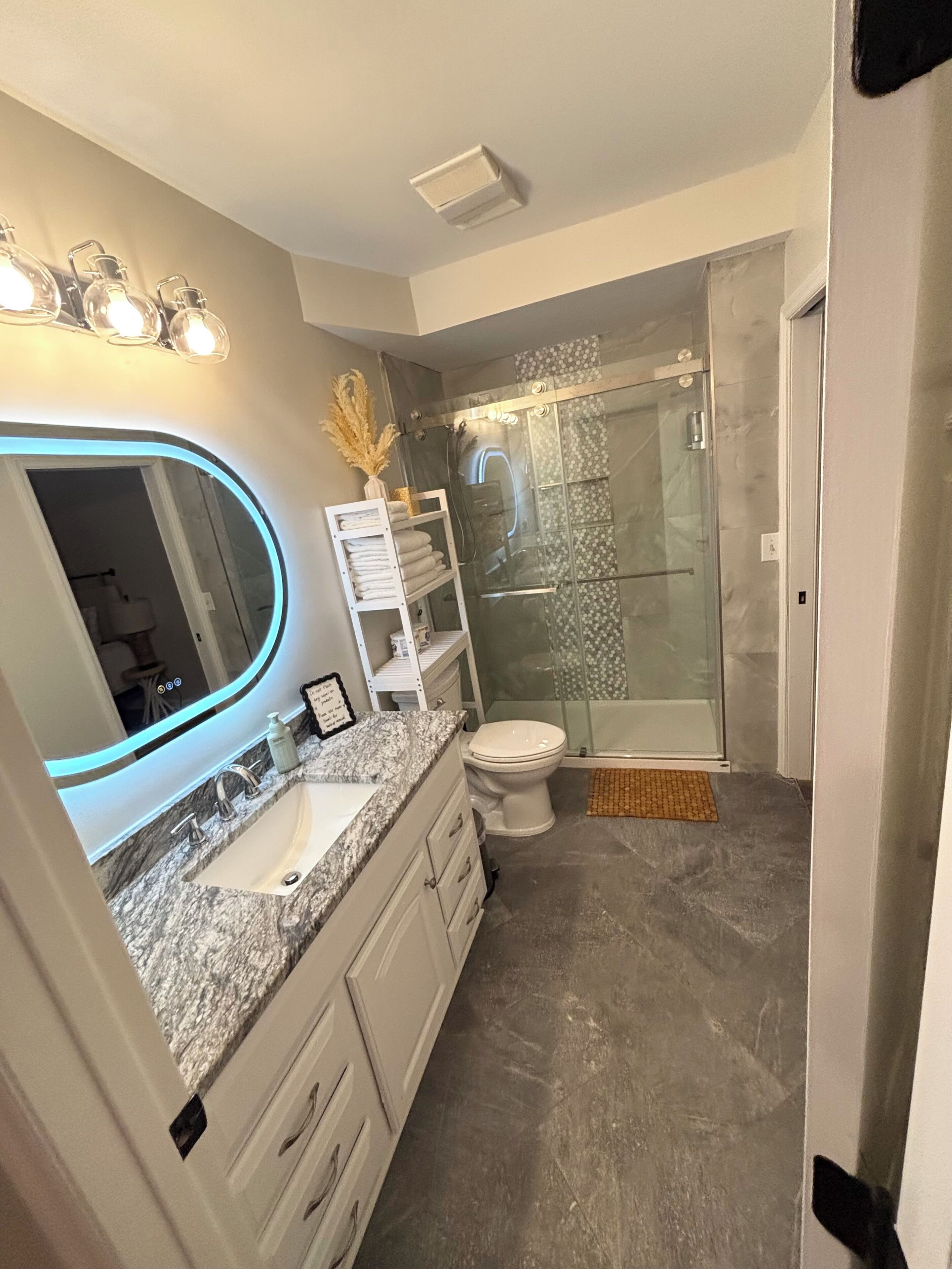 A modern bathroom with a granite countertop, a large mirror with under-cabinet lighting, a glass shower enclosure, and a white toilet.