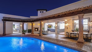 Ein Pool mit Pergola und Sitzbereich im Freien, verbunden mit einem Gebäude, das ein Feuerbecken und einen Essbereich bietet.