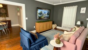A living room with a blue sofa, a pink sofa, a wooden cabinet, a dining area with a table and chairs, and a ceiling fan. - Boutique Historic Stay • Best Downtown Location (Savannah)
