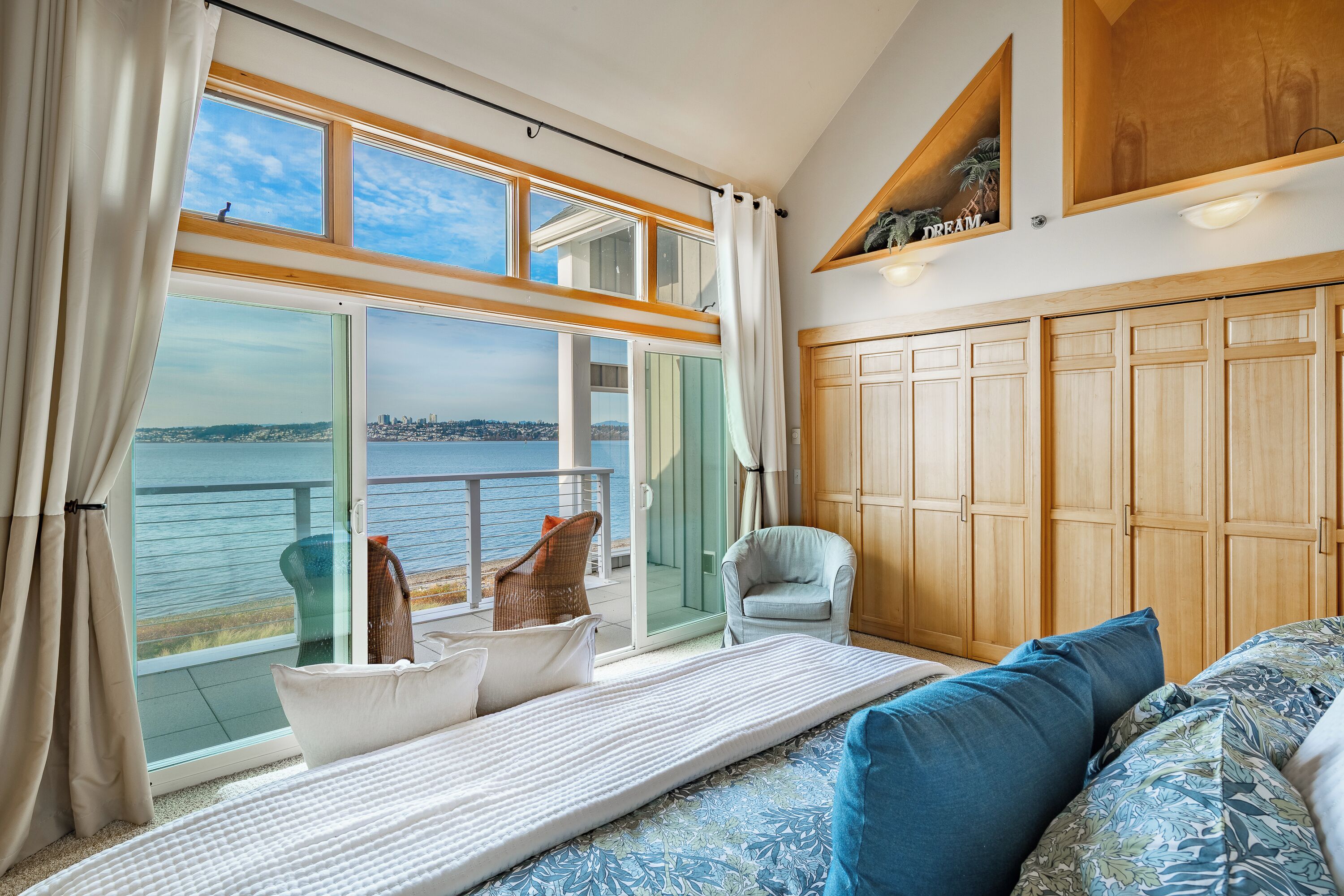 Ein Zimmer mit großem Fenster mit Blick auf ein Gewässer, einem Bett mit gemusterten Laken, einem Sessel und Holzschränken.