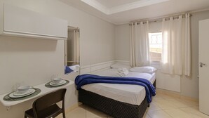 A bedroom with a bed, a desk with a chair, a mirror, and a window with curtains. - 05 Studio Prox Expo Berrini e Congonhas (São Paulo)