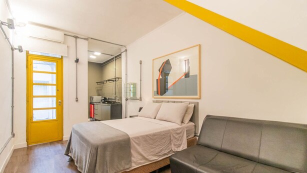 Un dormitorio con cama, sofá, una puerta amarilla y una cocina al fondo.