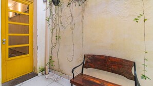 Una puerta amarilla con una ventana, un banco de madera y plantas colgantes frente a una pared con textura.