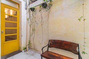 Una puerta amarilla con una ventana, un banco de madera y plantas colgantes frente a una pared con textura.