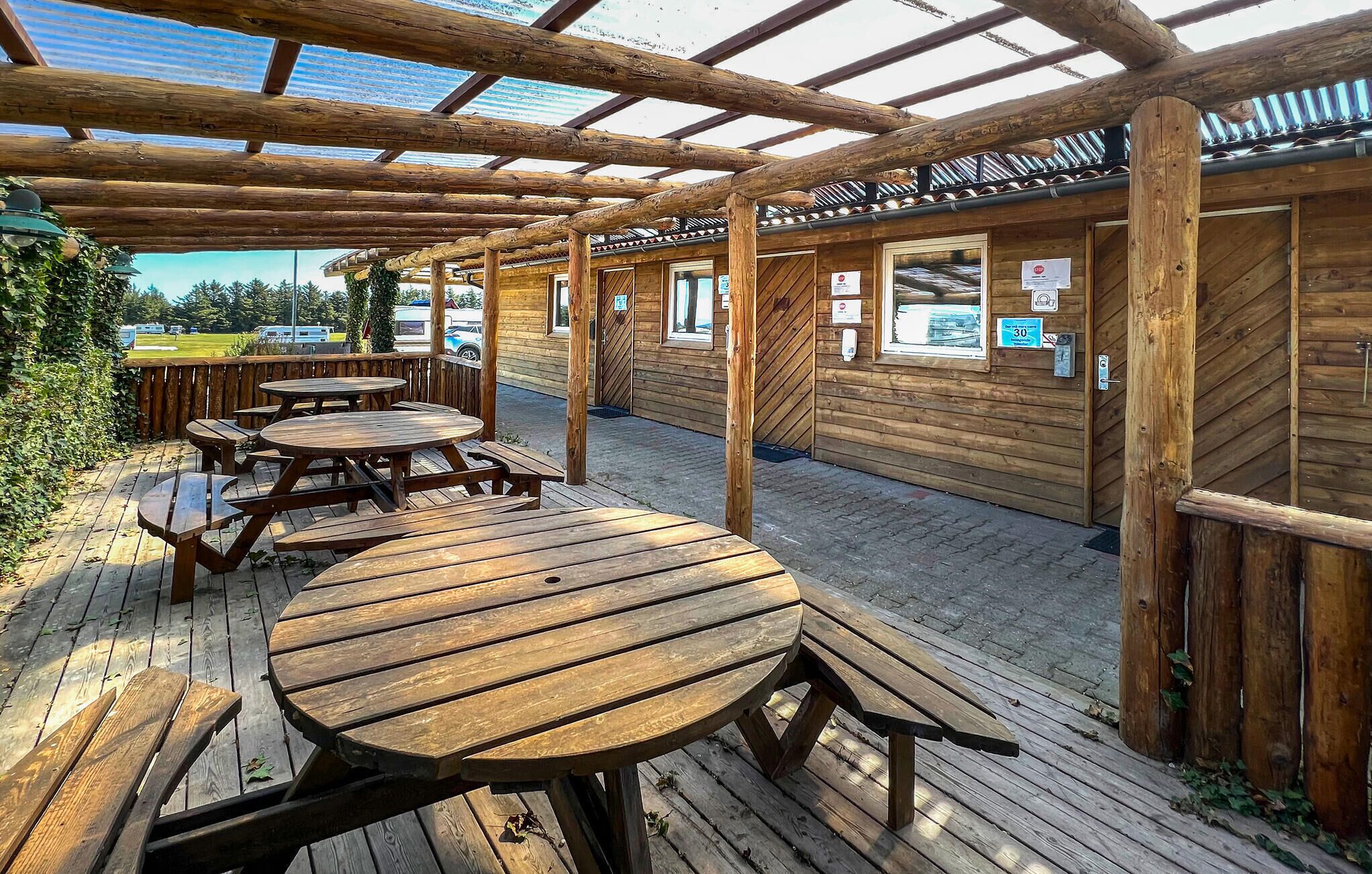 Una terraza de madera al aire libre con mesas de picnic y un área cubierta con vigas de madera y techo de metal corrugado.