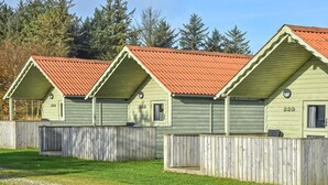 Une rangée de cabanes en bois aux toits rouges, numérotées 24 et 26, est entourée d’une clôture en bois et se dresse sur un fond de forêt.
