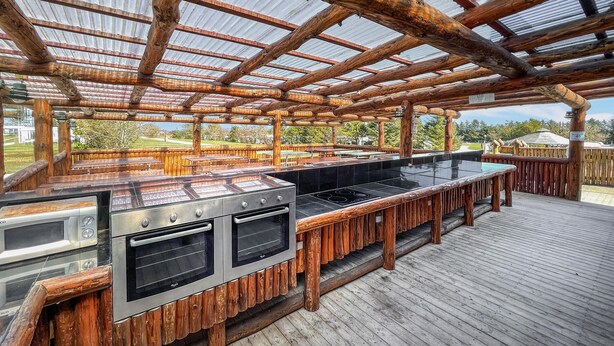 Ein Außenküchenbereich mit Holzterrasse, Metallgrill und einer Glasveranda.