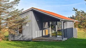 Een klein, gelijkvloers houten huisje met een rood dak, nummer 32, gelegen in een grasrijke omgeving met bomen.