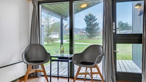 Een kamer met twee stoelen en een tafel, grote ramen met gordijnen, en uitzicht op een gazon en bomen.