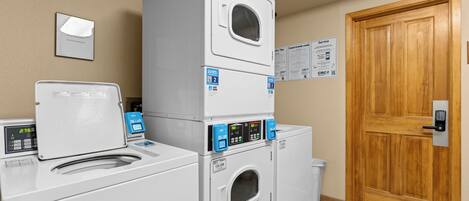 Une buanderie équipée de machines à laver et de sèche-linge superposés, une porte en bois et des panneaux d’information fixés au mur.