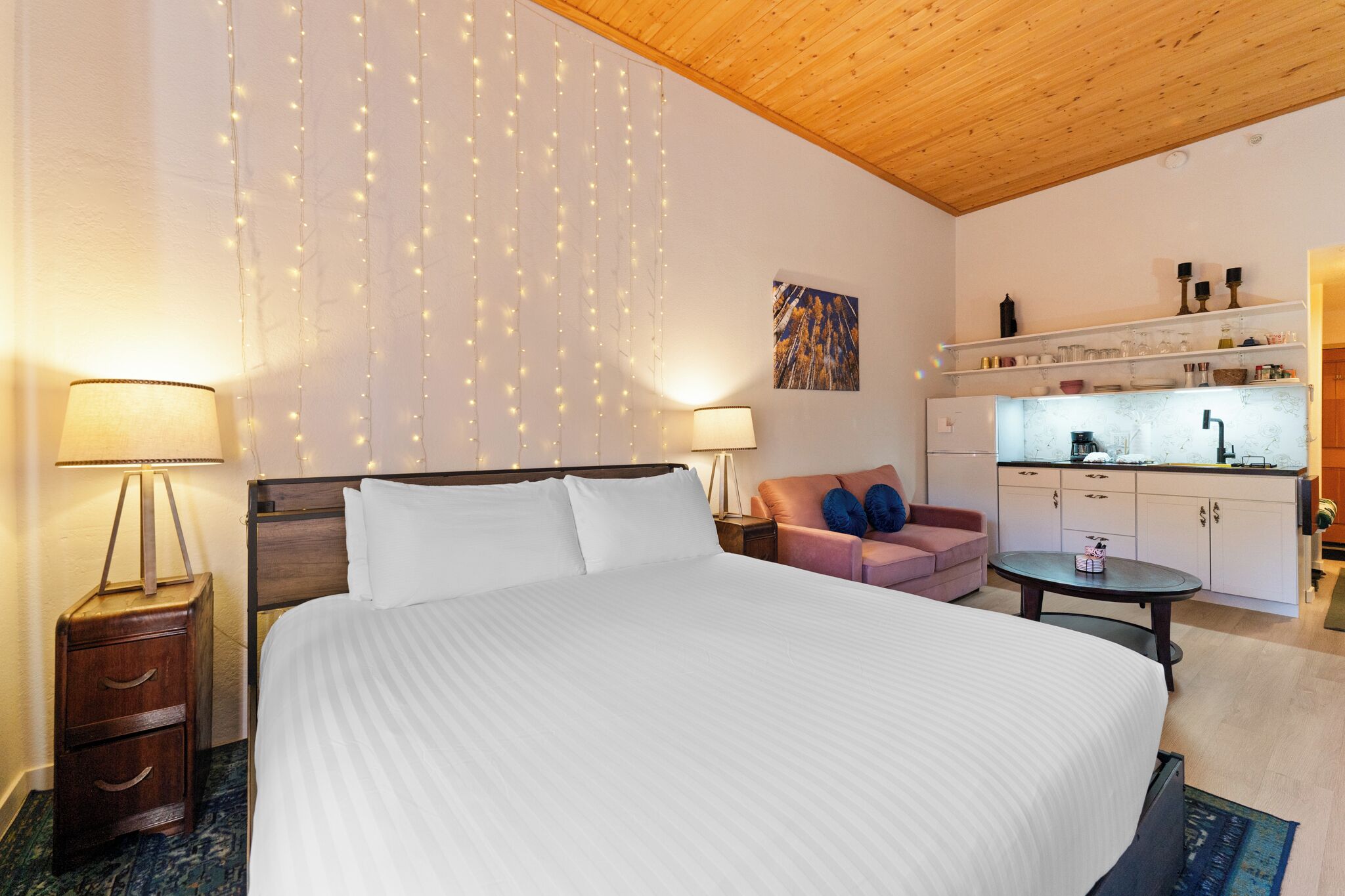 A hotel room with a large bed, bedside lamps, a sofa, a small table, and a kitchenette.