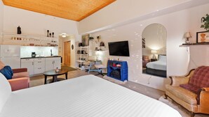 Quarto de hotel moderno com teto de madeira, cozinha, TV fixada na parede e um espaço de estar acolhedor com lareira.