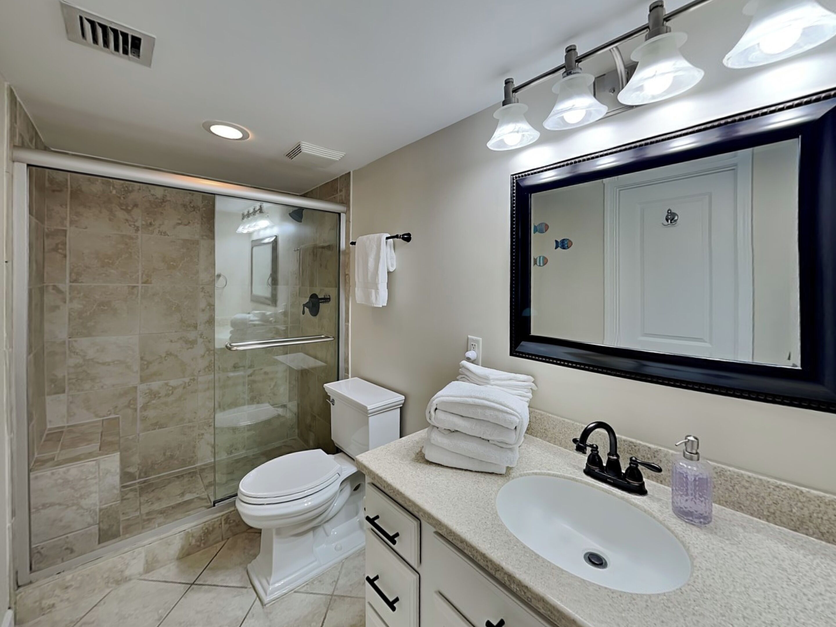 A modern bathroom with a glass-enclosed shower, a white toilet, a sink with a soap dispenser, and a large mirror.