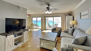 A spacious living room with a flat-screen TV, a ceiling fan, and a view of the ocean through large sliding doors. - Regency Towers #401 East - Sea La Vie (Pensacola Beach)