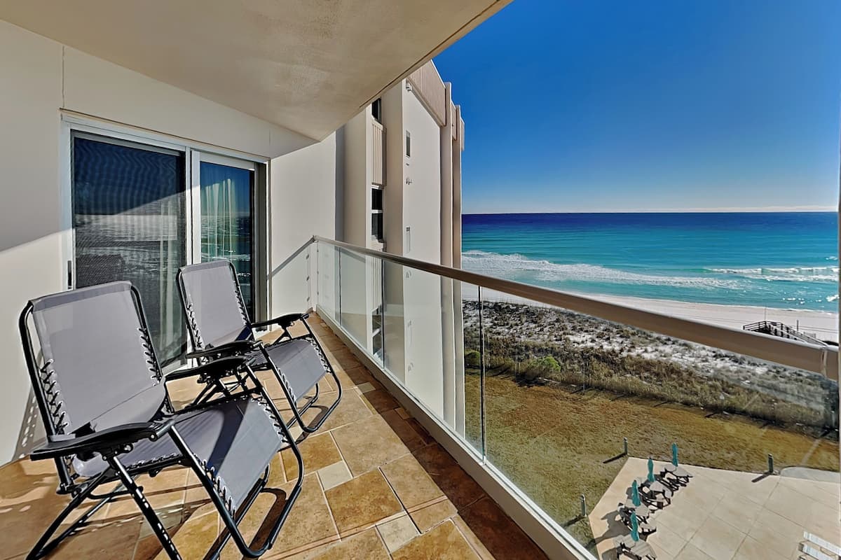 Varanda com duas cadeiras e uma mesa, com vista para uma praia de águas azuis cristalinas e costa rochosa.