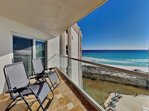 Ein Balkon mit zwei Stühlen und einem Tisch, mit Blick auf einen Strand mit klarem, blauem Wasser und einer felsigen Küste.