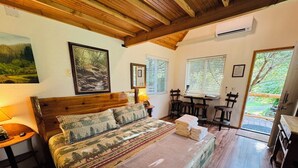 Une chambre à coucher avec une tête de lit en bois, un lit recouvert d’une literie à motifs, une table de chevet avec une lampe, et une vue sur l’extérieur par une porte.