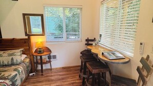 A cozy room with a wooden ceiling, a bed with a forest-themed bedspread, a small round table with a lamp, and a dining area with wooden chairs.