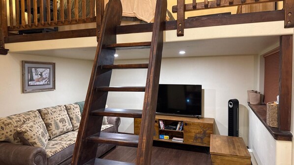 A loft-style room with a wooden ladder leading to a bunk bed, a flat-screen TV, a sofa, and a small wooden shelf with books and decorative items. - Cozy Mountain Loft Retreat in Stunning Paradise Valley (Emigrant)