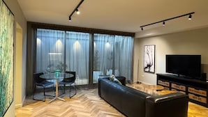 Ett modernt vardagsrum med ett fiskbensmönstrat trägolv, en svart lädersoffa, ett matbord i glas med stolar, en platt-TV och ett stort fönster med tunna, genomskinliga gardiner.