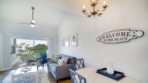 Una sala de estar moderna con un ventilador de techo, una araña, un cartel que dice 'Welcome to the Beach', un comedor con una mesa y sillas, y un sofá con vistas al exterior.