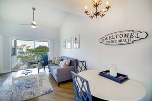 Ein modernes Wohnzimmer mit Deckenventilator, einer Kristallleuchter, einem Schild mit der Aufschrift 'Welcome to the Beach', einem Essbereich mit Tisch und Stühlen sowie einer Couch mit Blick ins Freie.
