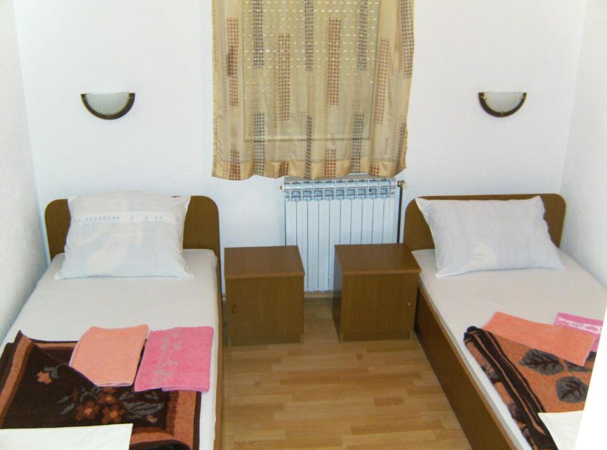 Una habitación pequeña y sencilla con dos camas, un radiador y una ventana con cortina.