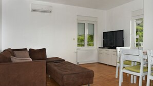 Een woonkamer met een bruine bank, een witte eettafel met stoelen, een flatscreen televisie en een raam met jaloezieën.