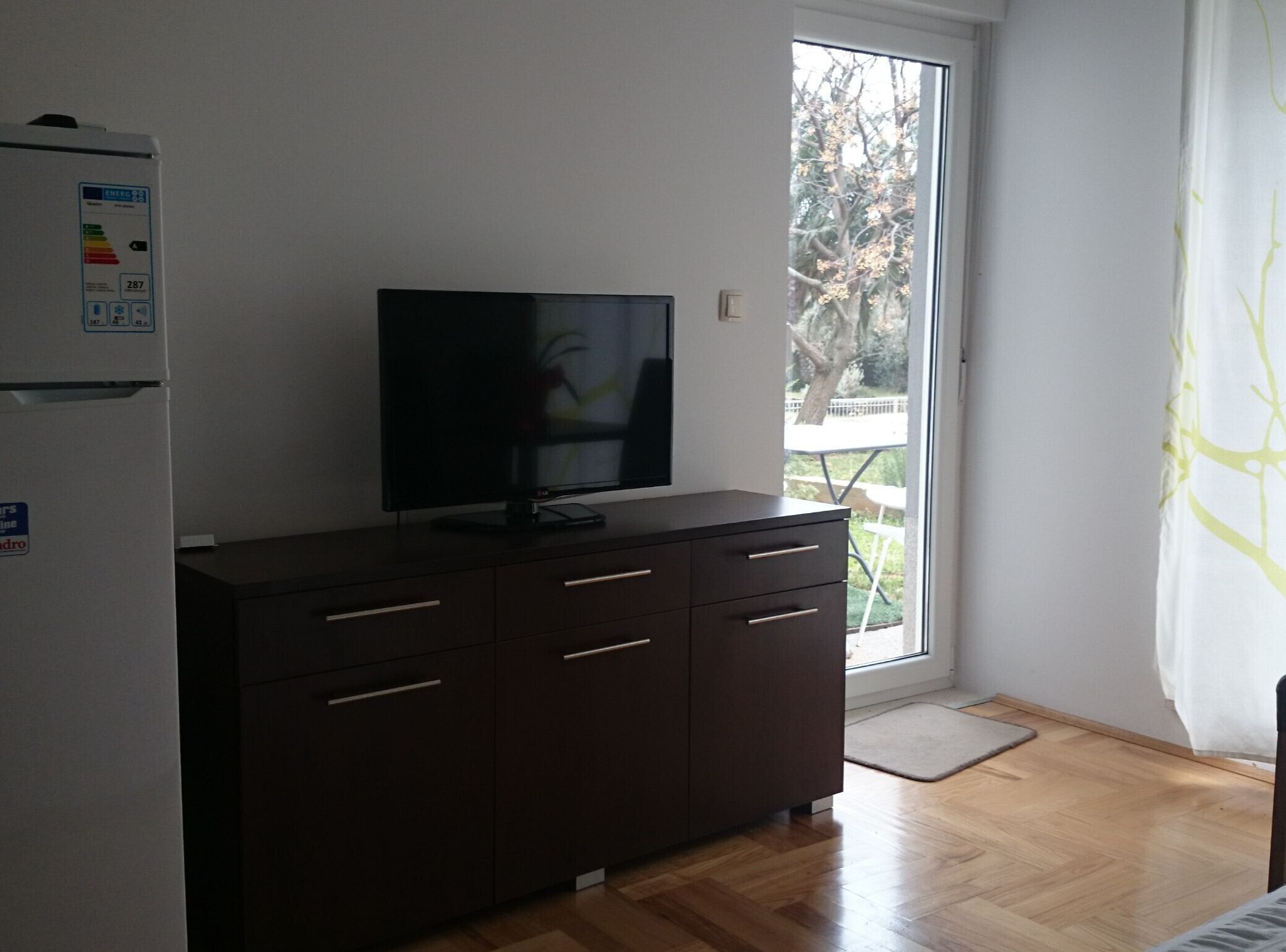 Habitació amb un armari de fusta fosca, un televisor de pantalla plana a la part superior, un frigorífic blanc i una porta corredissa de vidre que dona a una zona exterior amb taula i cadires.