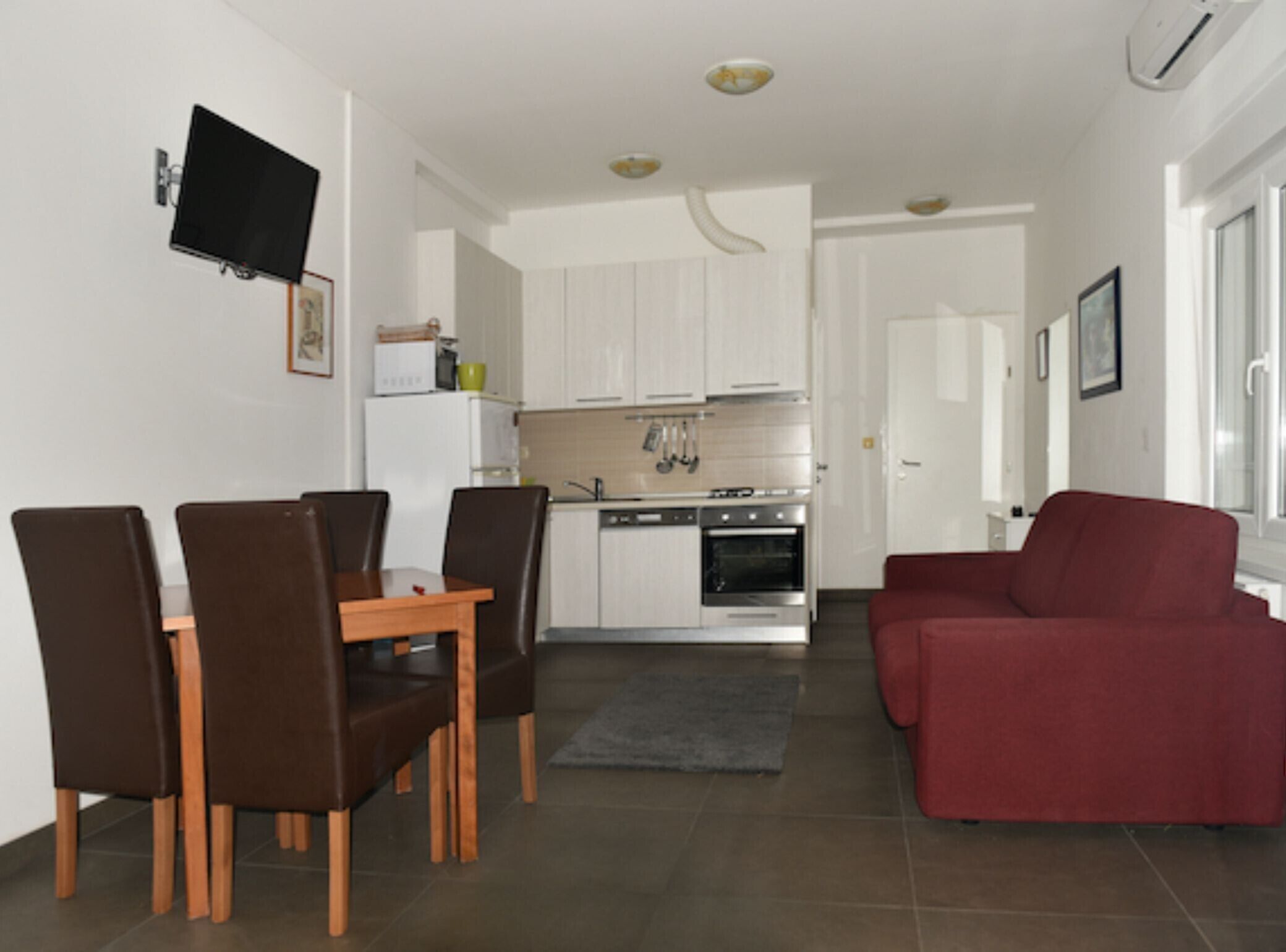 Ein modernes Wohnzimmer mit einem roten Sofa, einem Esstisch mit Stühlen, einer Küchenzeile mit weißen Schränken und einem flachbildfernseher an der Wand.