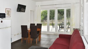 Una sala de estar con un sofá rojo, una mesa de comedor con sillas, un televisor montado en la pared y una puerta corrediza de vidrio que da a un espacio exterior.
