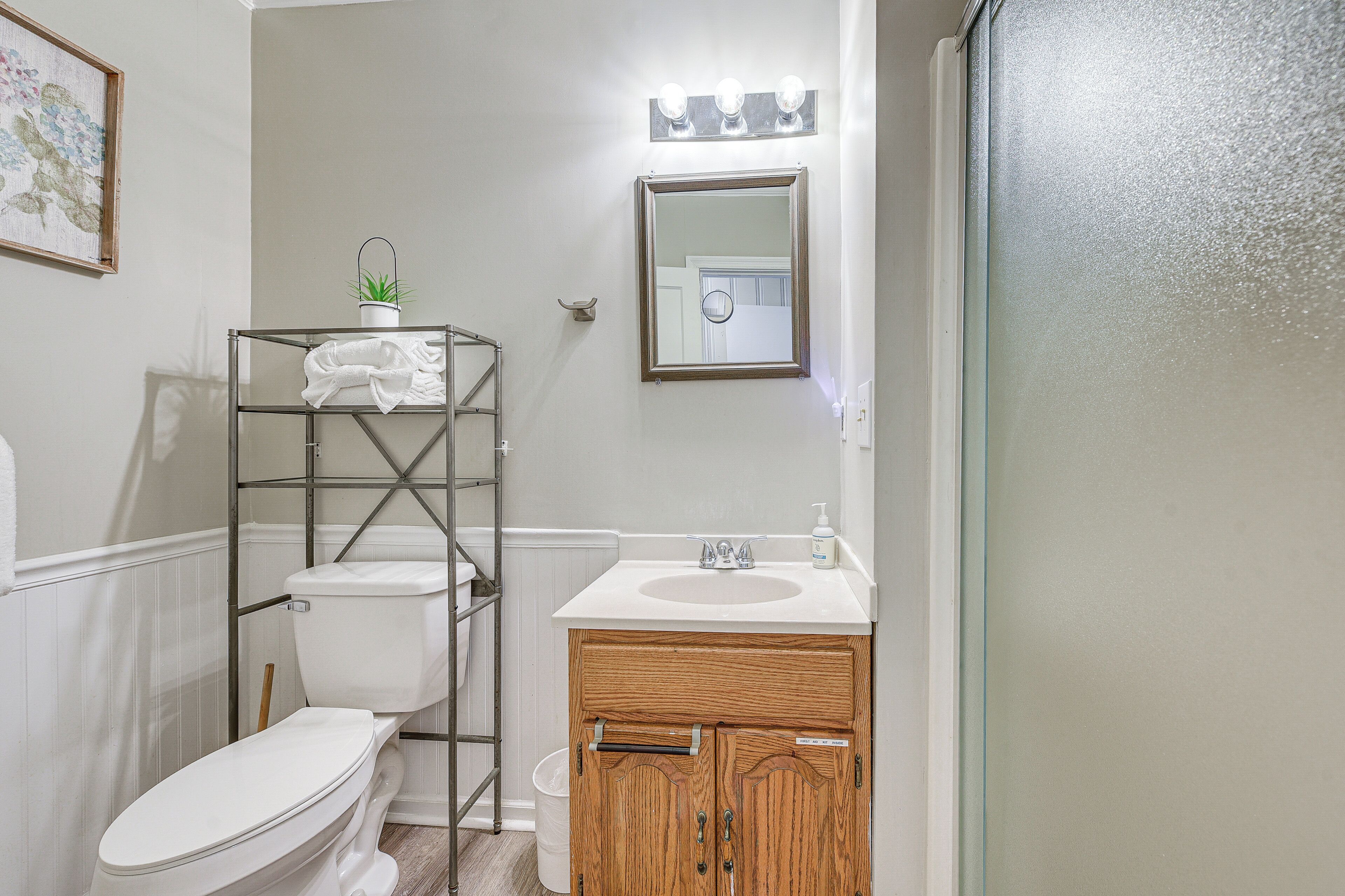 Une salle de bain avec des toilettes blanches, un meuble-lavabo en bois et une étagère métallique sur laquelle est posée une plante.
