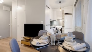 Uma área de refeições moderna com uma mesa redonda posta para quatro pessoas, uma televisão e uma cozinha ao fundo.