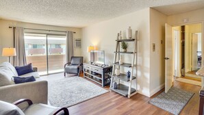 A living room with a sofa, a coffee table, a TV, a bookshelf, and a door leading to another room. - Steps to CSULB: Cozy Condo w/ Pool Access! (Long Beach)