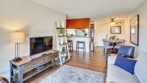 A modern living room with a flat-screen TV, wooden entertainment unit, and a sofa with a blue pillow. A kitchen is visible in the background with wooden cabinets and a dining area. - Steps to CSULB: Cozy Condo w/ Pool Access! (Long Beach)