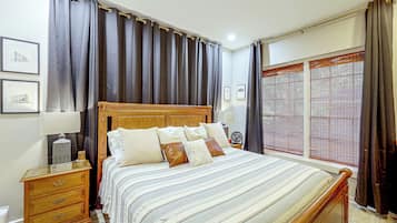 A bedroom with a wooden bed, a nightstand, a lamp, a bedside table with a candle, and a window with blinds.