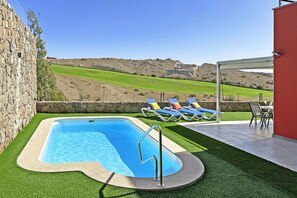 A swimming pool with artificial grass, a stone wall, and a view of a green field and hills. - Salobre Vista Golf 24 (Maspalomas)