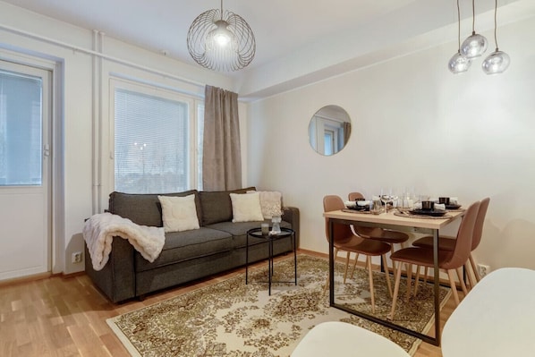 A modern living room with a sofa, dining table, chairs, and a coffee table. There is a large window with blinds, a ceiling light fixture, and a mirror on the wall. - Arctos City Suite, private sauna By Lapland Host (Rovaniemi)